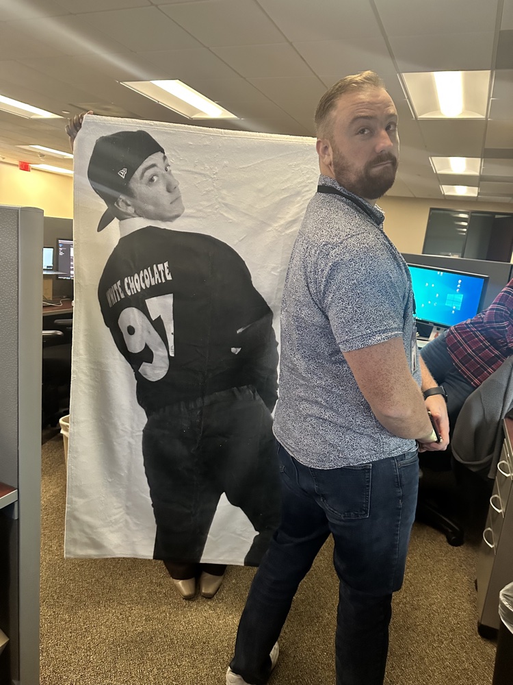 Daniel at the office next to a blanket printed with his teenage dance photo, jersey reading White Chocolate